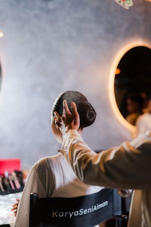 Hair stylist applying finishing touches to an elegant updo bun hairstyle at Karya Seni Aiman salon with round mirror.