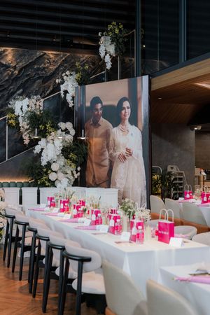 Elegant wedding reception venue with long white tables, pink accents, floral arrangements, and a large couple portrait backdrop.