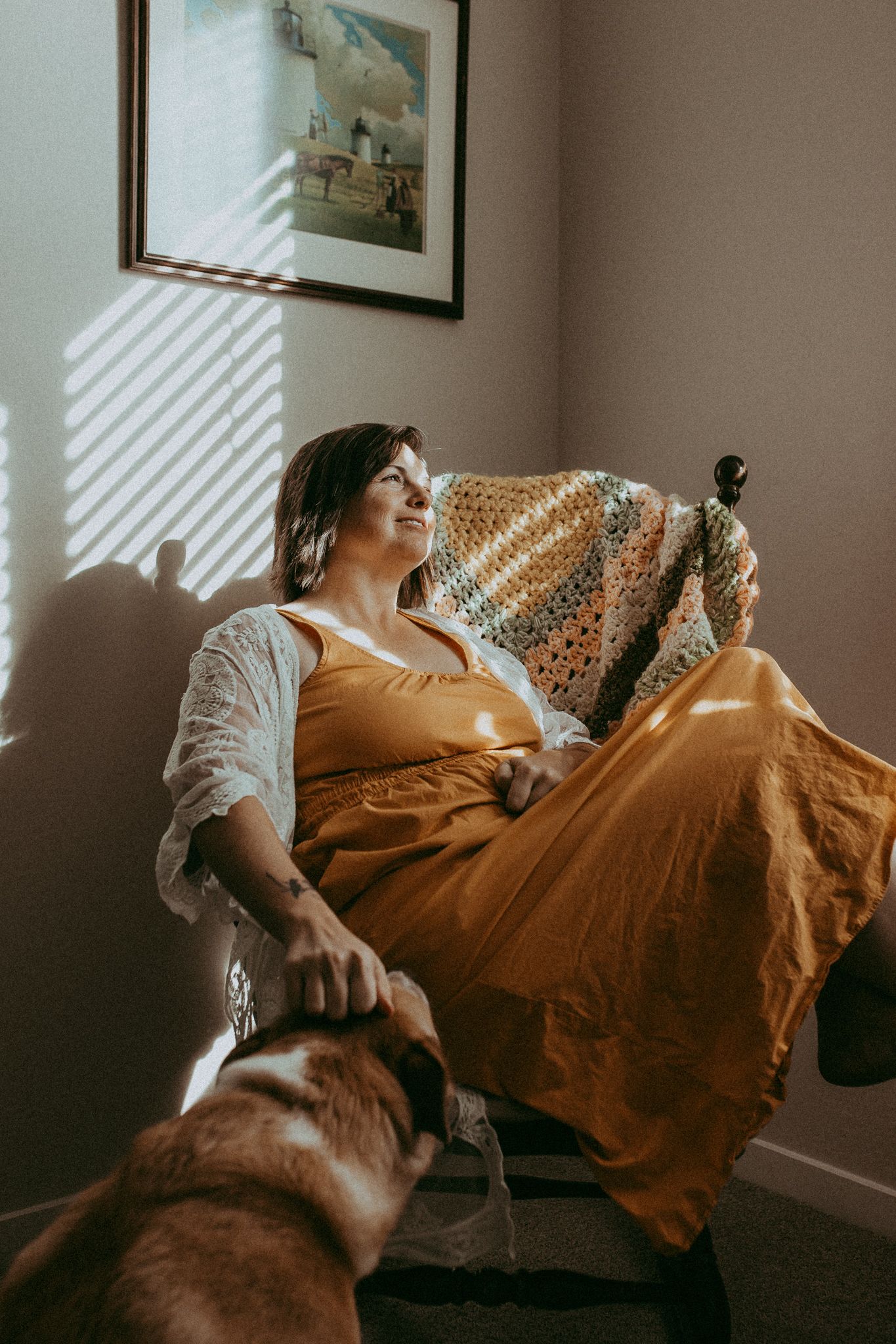 Woman in mustard dress relaxing in chair with dog, bathed in warm sunlight beside a framed wall art.