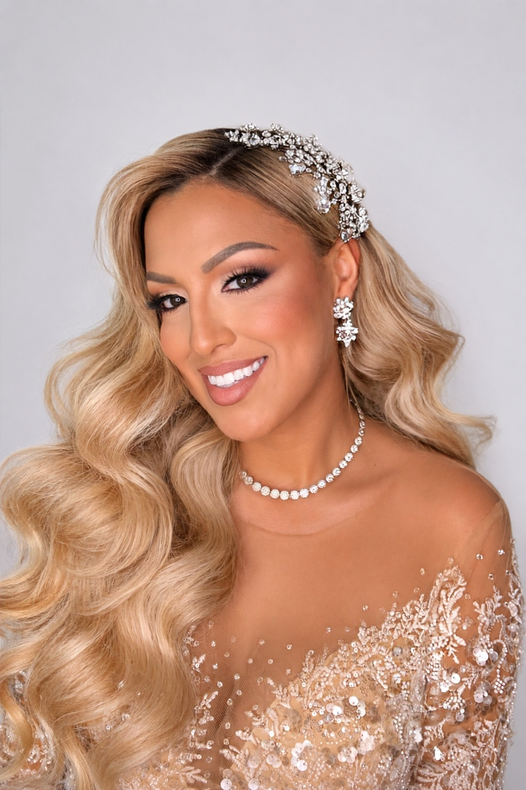 Elegant woman wearing crystal hair accessory, pearl necklace, and beaded off-shoulder gown with glamorous wavy blonde hair.