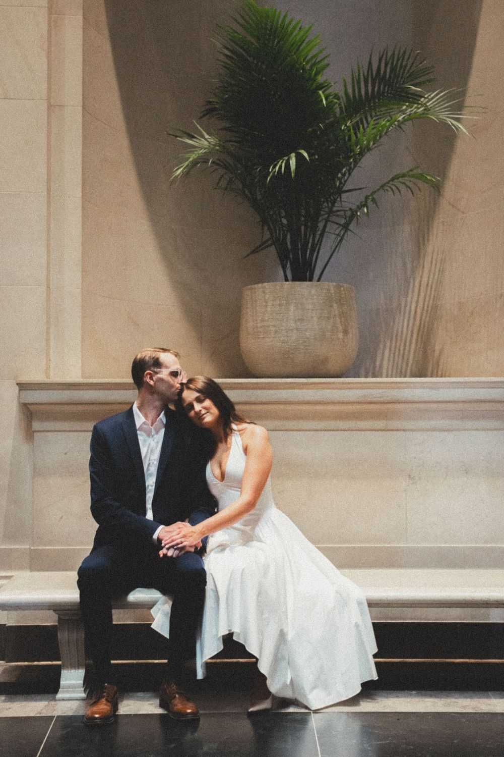 Couple in wedding attire sitting on a bench, groom kissing bride's forehead, with a palm plant backdrop.