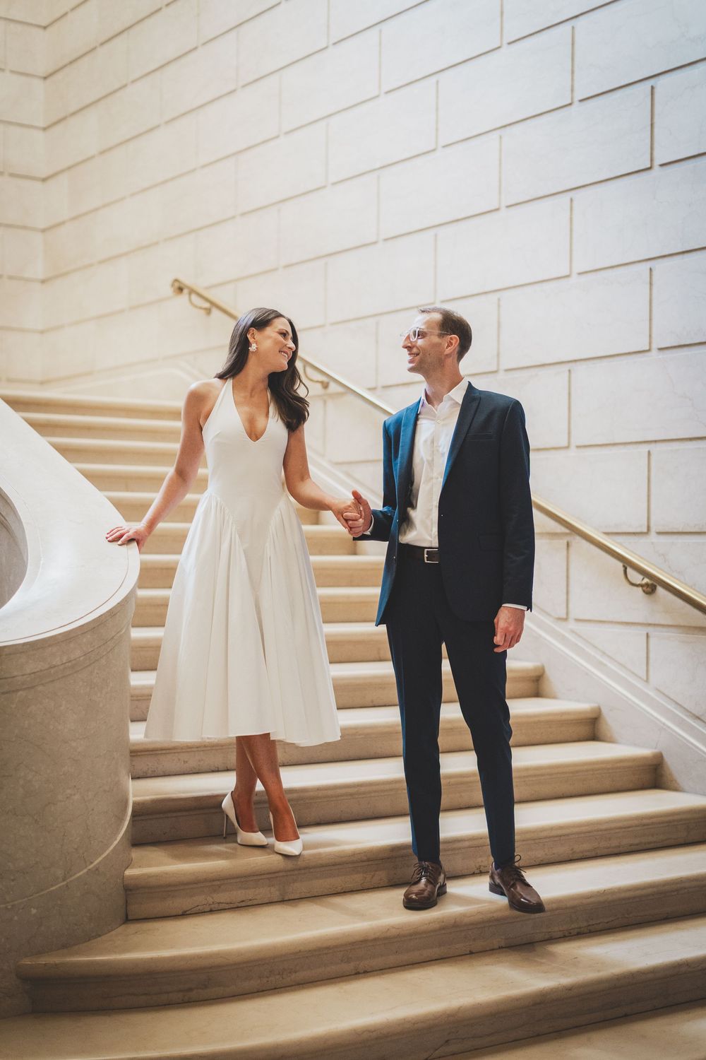 Elegant couple holding hands on marble stairs, woman in white midi dress and man in navy suit, in a grand building.