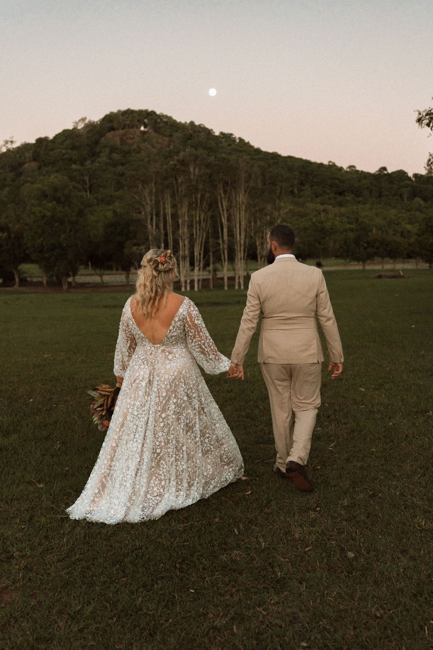 Bride and groom walking hand in hand across green lawn at dusk with moonrise over lush hillside backdrop.