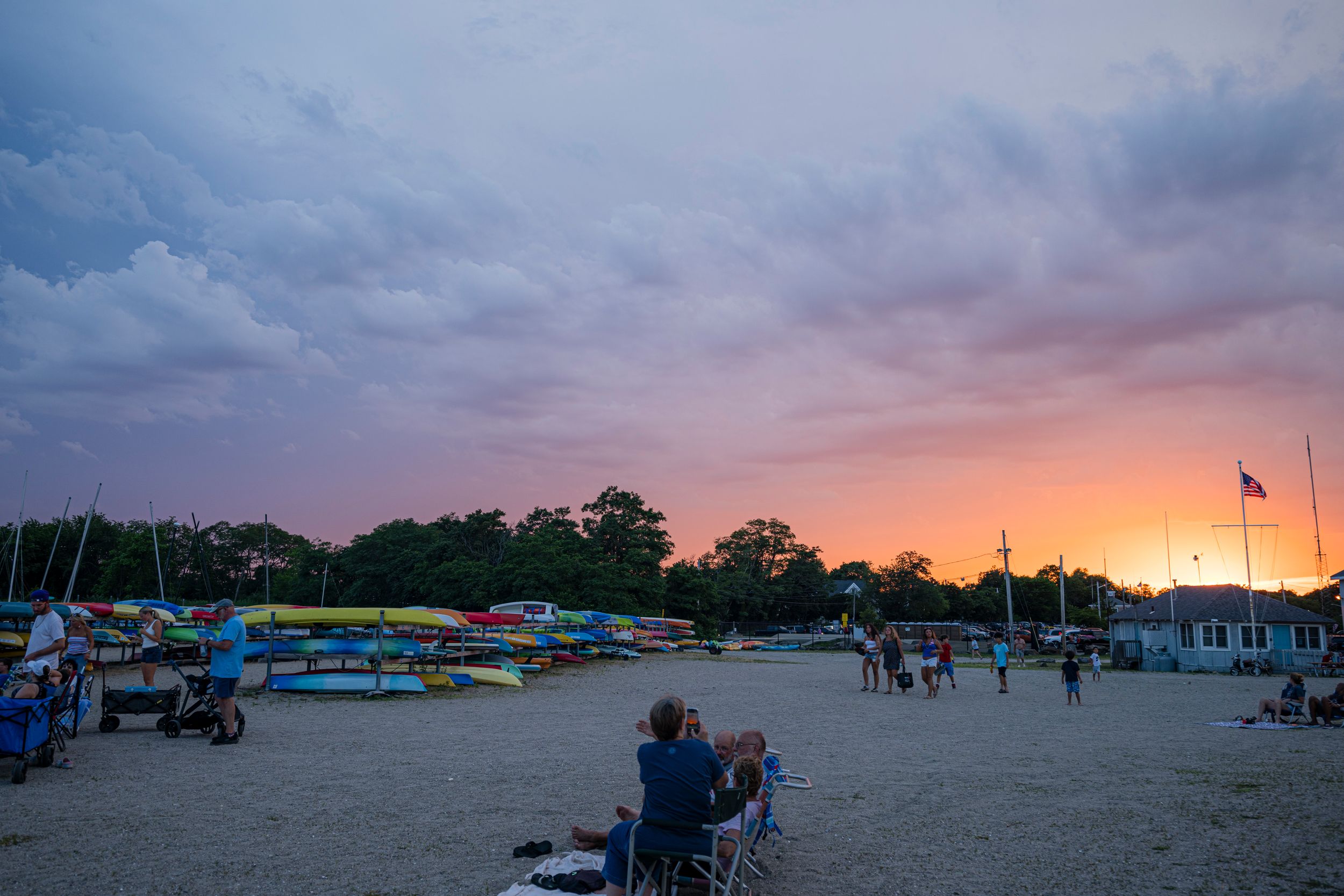 People relax on a sandy beach at sunset with colorful boats and dramatic cloudy skies in the background.