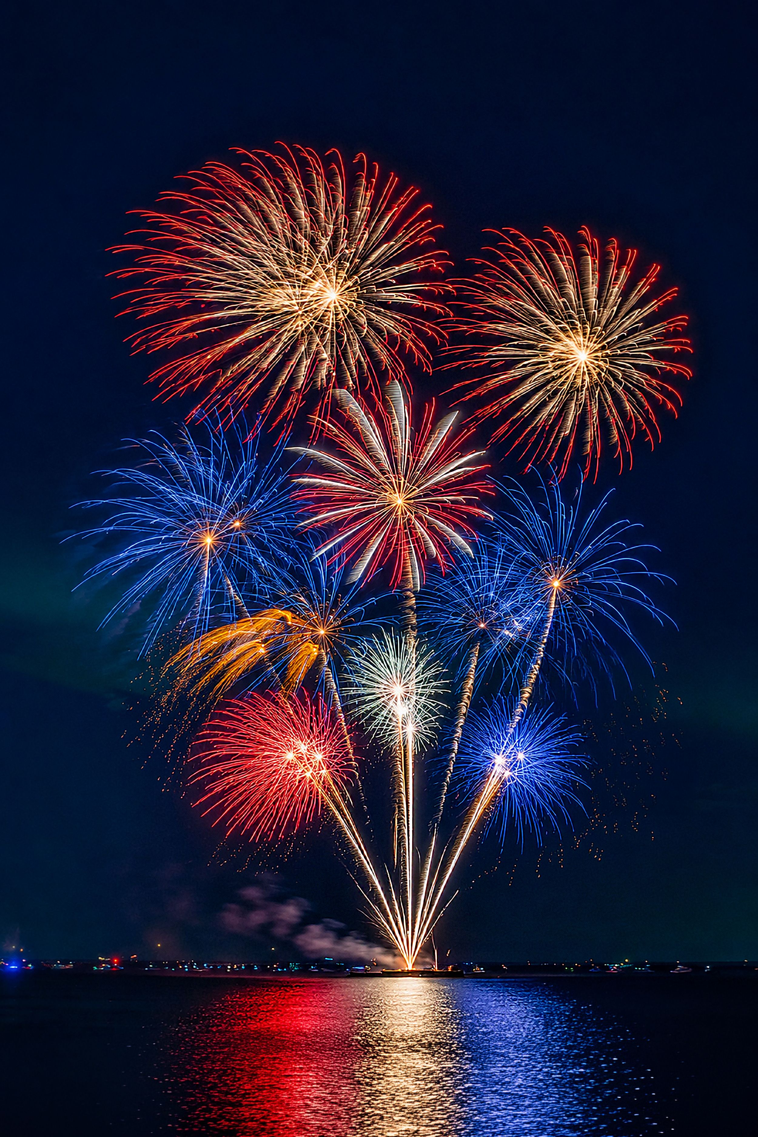 Colorful red, white, and blue Fourth of July fireworks exploding over water with vivid reflections at night.