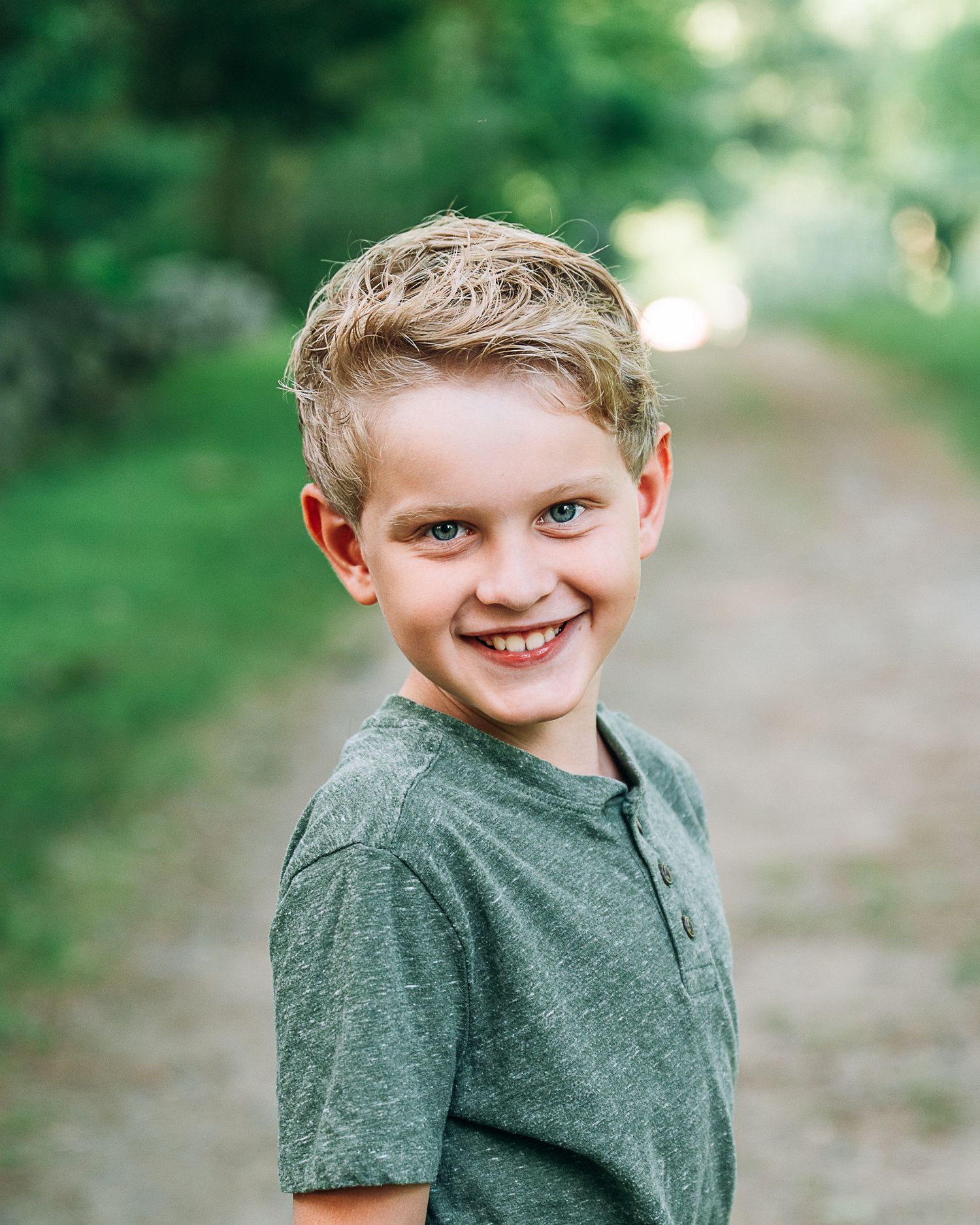Smiling young boy with blonde hair wearing a green t-shirt, posing outdoors on a tree-lined path.