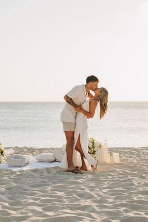 Couple kissing on a sandy beach at sunset, surrounded by white flowers and candles in a romantic proposal setup.
