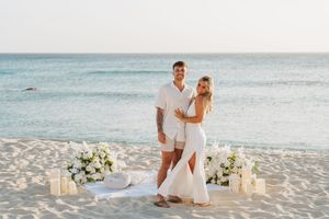 Couple in white outfits posing on a sandy beach with floral arrangements and candles by the ocean.