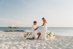 Man proposes to woman on white sand beach at sunset with floral arrangements and candles, sailboat in background.