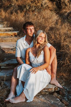 Couple in white outfits sitting on stone steps surrounded by golden dry grass in warm sunset light.