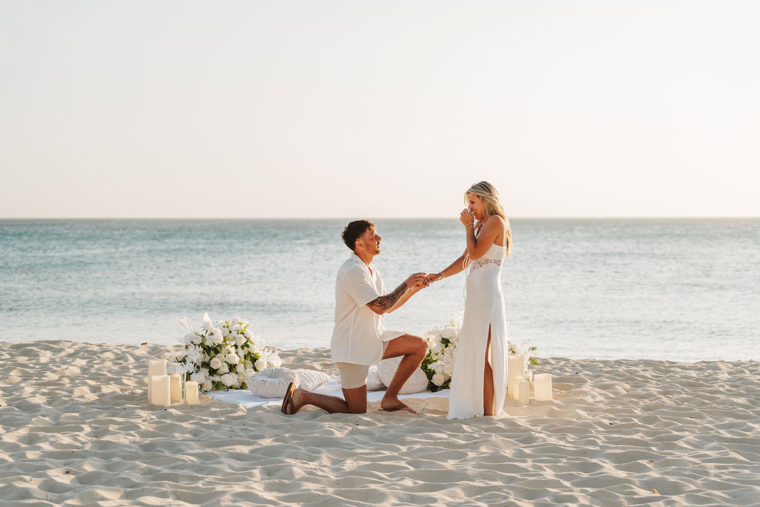 Man proposes to woman on a sandy beach at sunset, surrounded by white flowers and candles near the ocean.