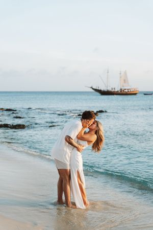 Romantic couple kissing on a tropical beach in white outfits with a sailing ship in the background.
