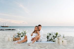 Couple in white attire sitting on beach with white floral arrangements and candles at sunset.