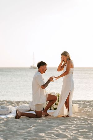 Man proposing to woman on sandy beach at sunset, both dressed in white, with ocean in background.