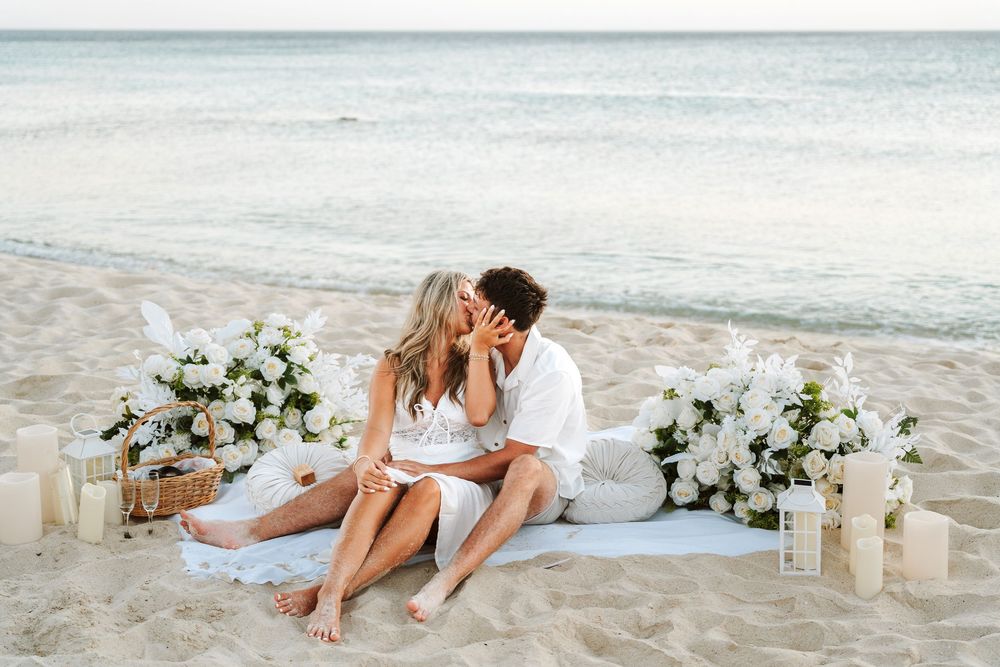 Couple sharing a romantic kiss on a white sand beach surrounded by white floral arrangements and candles.