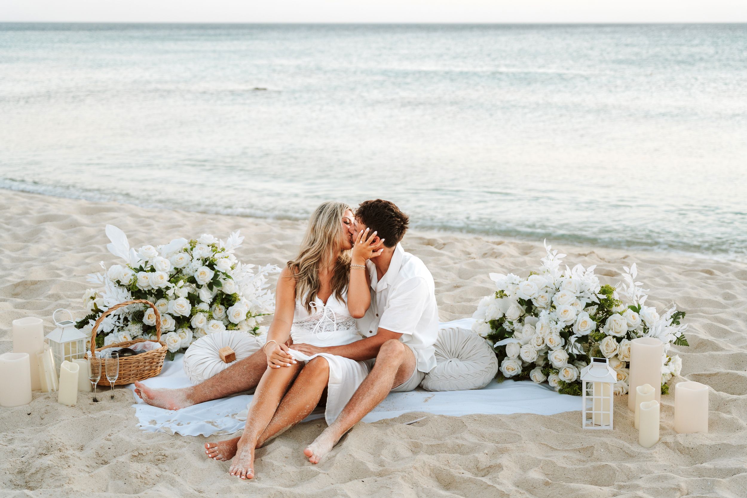 Couple sharing a romantic kiss on a white sand beach surrounded by white floral arrangements and candles.