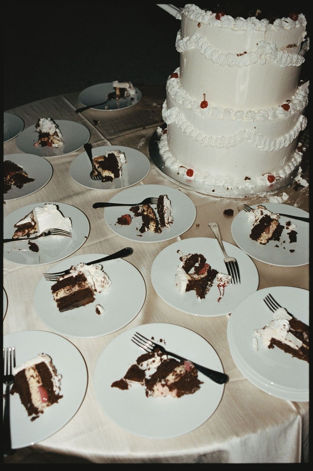 White two-tier wedding cake with cherry decorations surrounded by plated chocolate cake slices with forks on a reception table.
