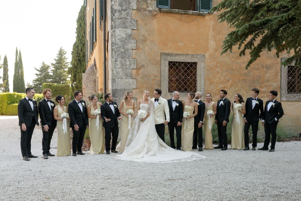 Wedding party poses outside a Tuscan villa, bridesmaids in sage gowns and groomsmen in black tuxedos flanking the bride and groom.