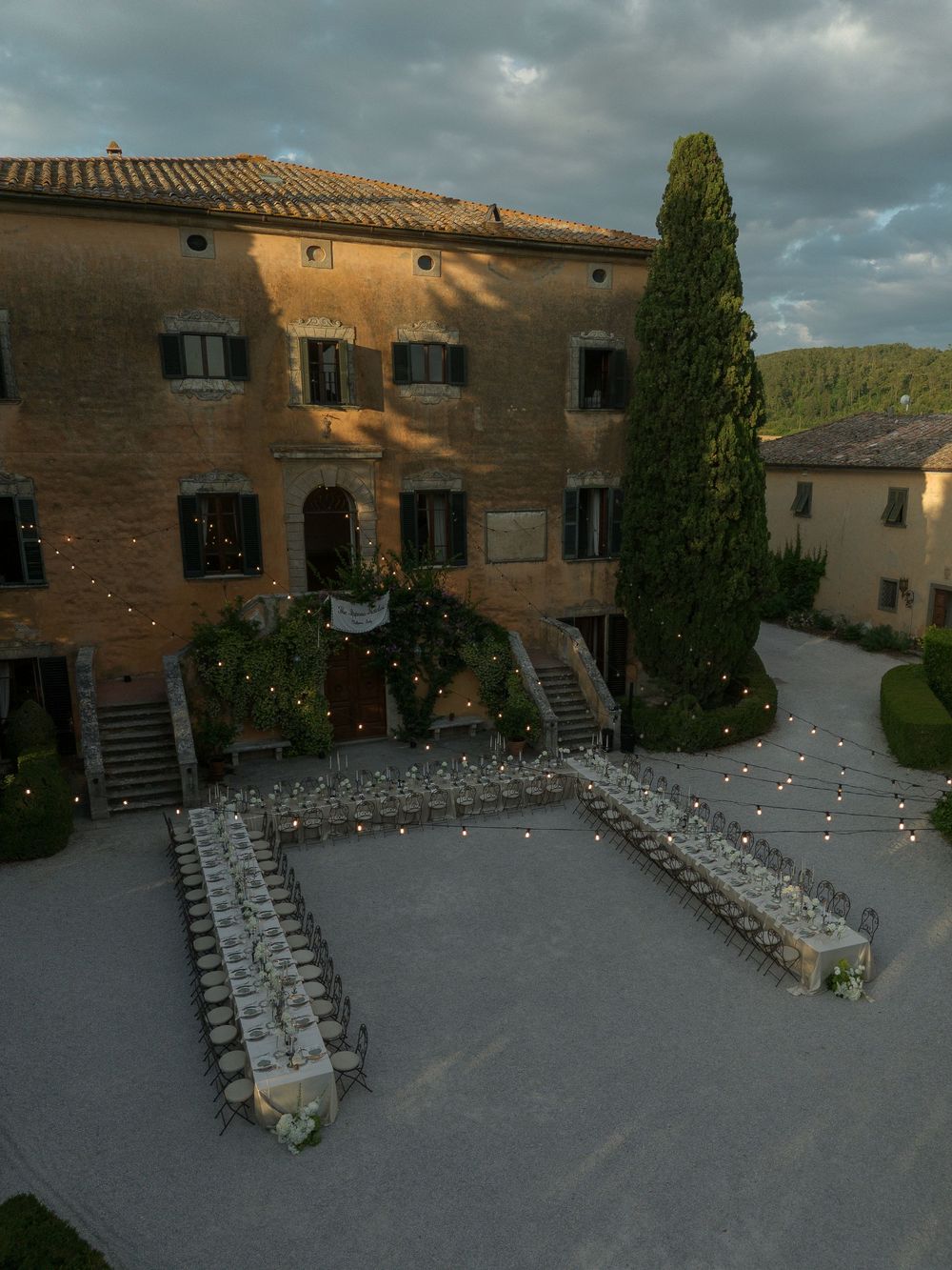 Aerial view of elegant outdoor wedding reception with long tables and string lights at a Tuscan villa at dusk.