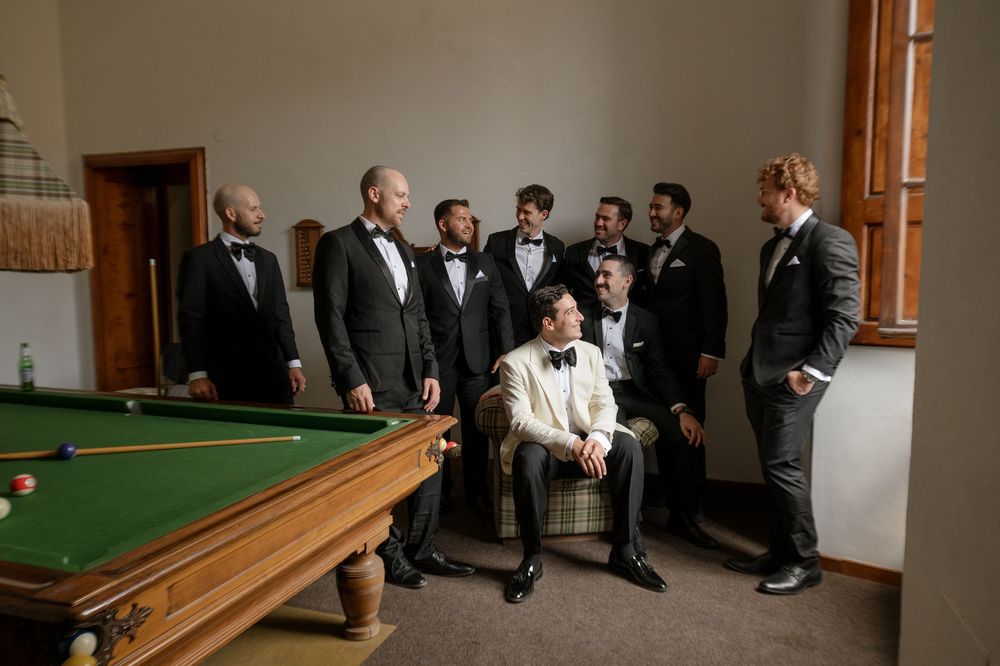 Groom seated in white suit surrounded by groomsmen in black tuxedos near a pool table in an elegant room.
