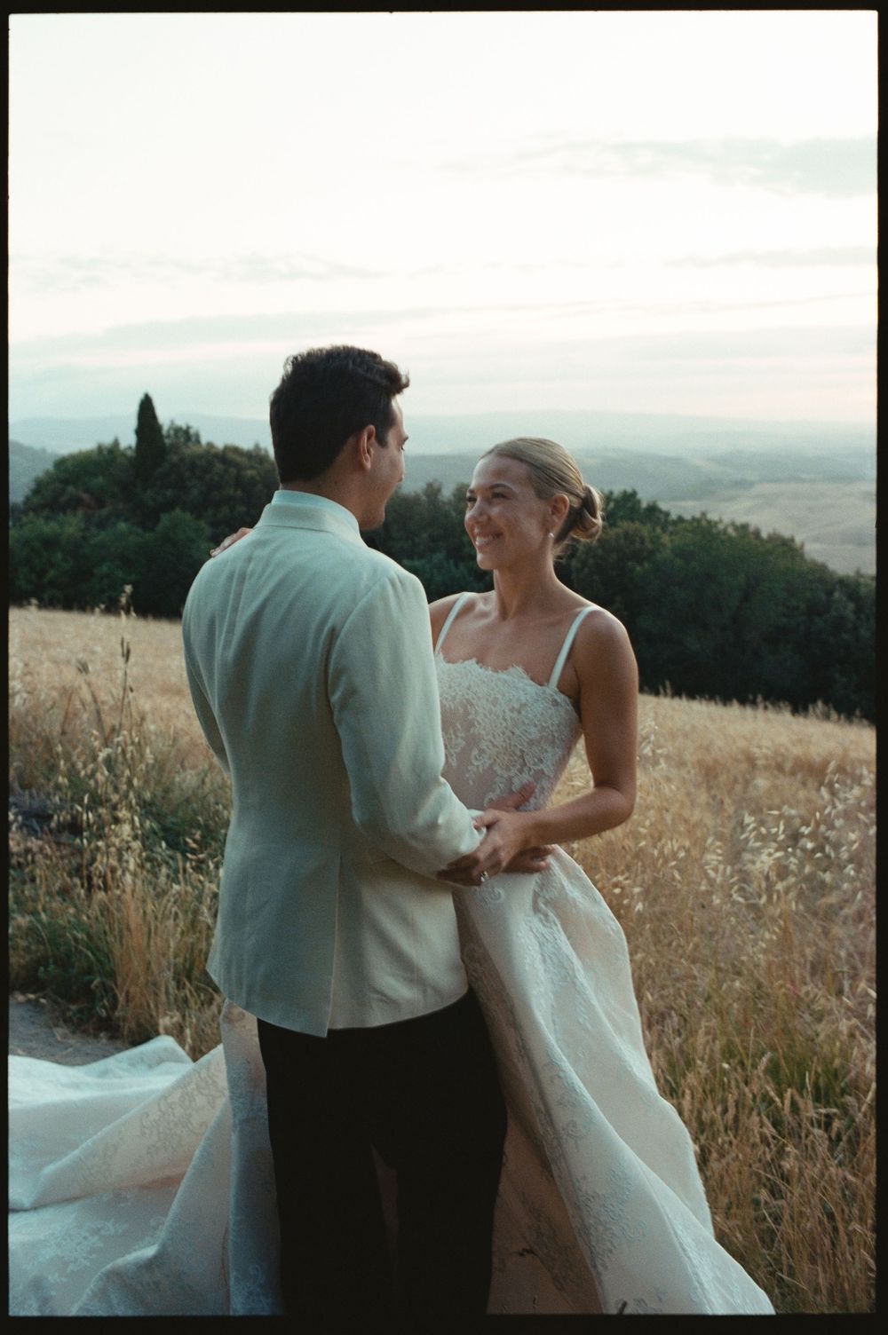 Bride and groom holding hands in a golden field at sunset with scenic Tuscan hills in the background.