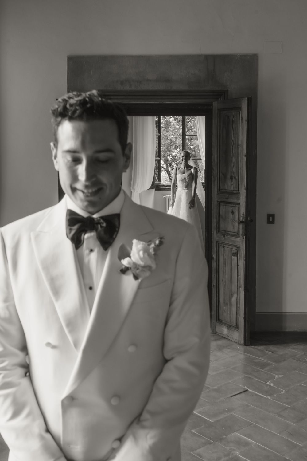 Black and white portrait of smiling groom in white tuxedo with bow tie and boutonniere inside a rustic villa.