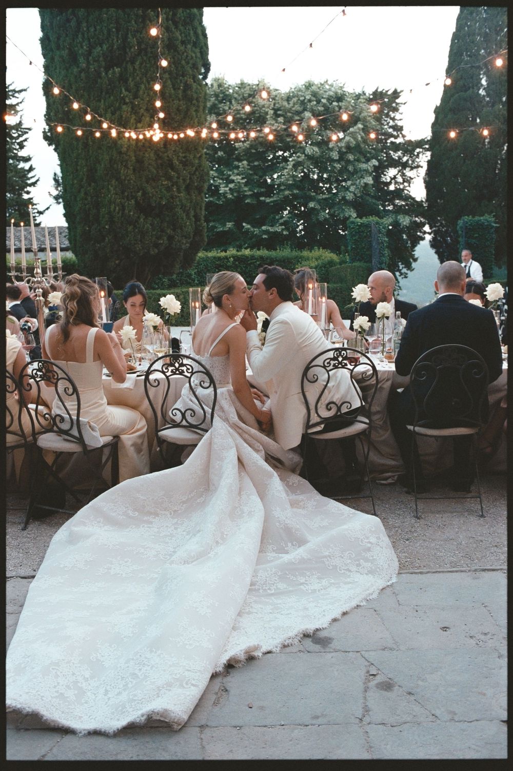 Bride and groom sharing a kiss at outdoor wedding reception with string lights and cypress trees in elegant garden setting.
