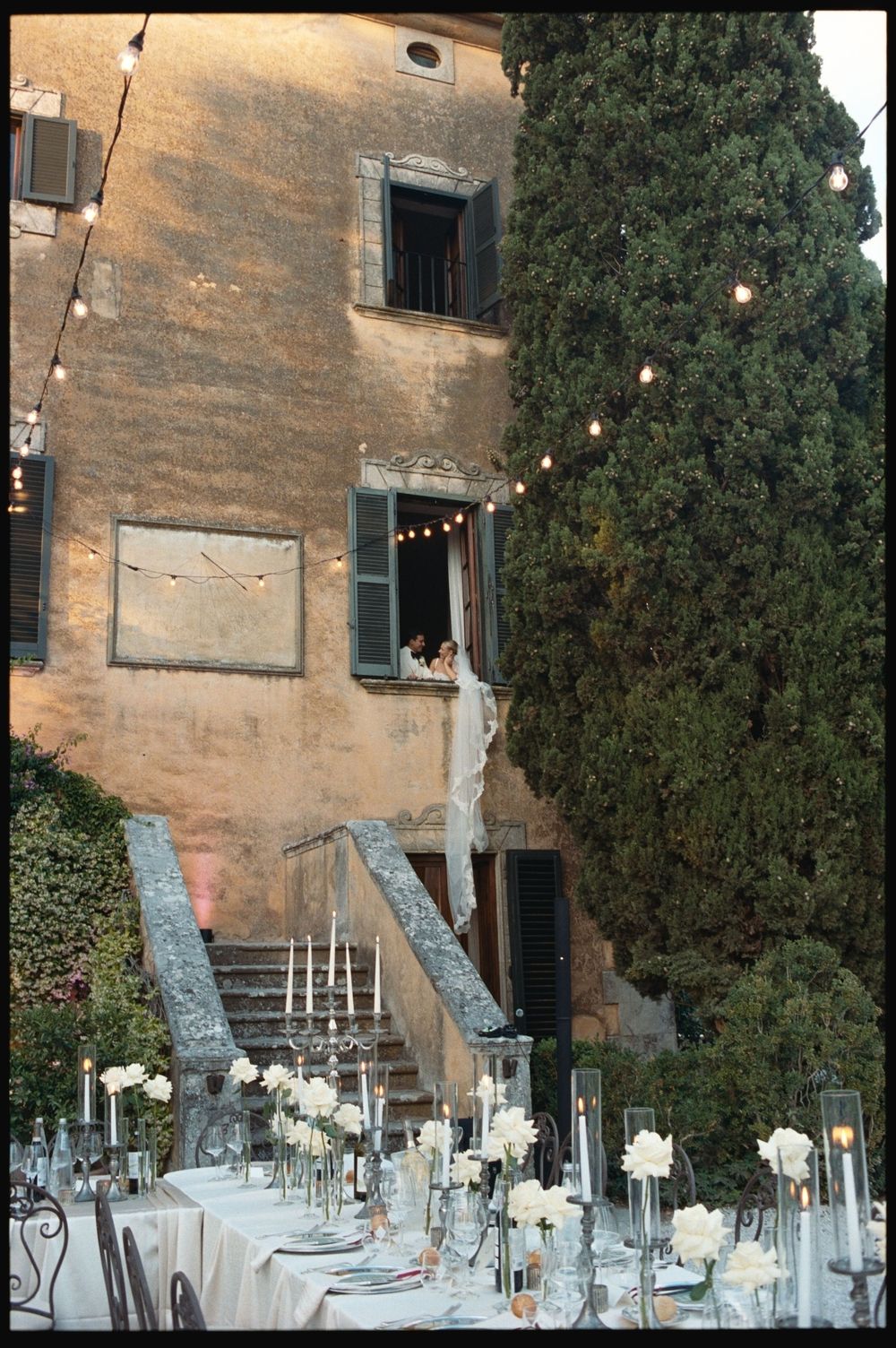 Elegant outdoor wedding reception at a rustic Italian villa with white floral centerpieces and string lights at dusk.