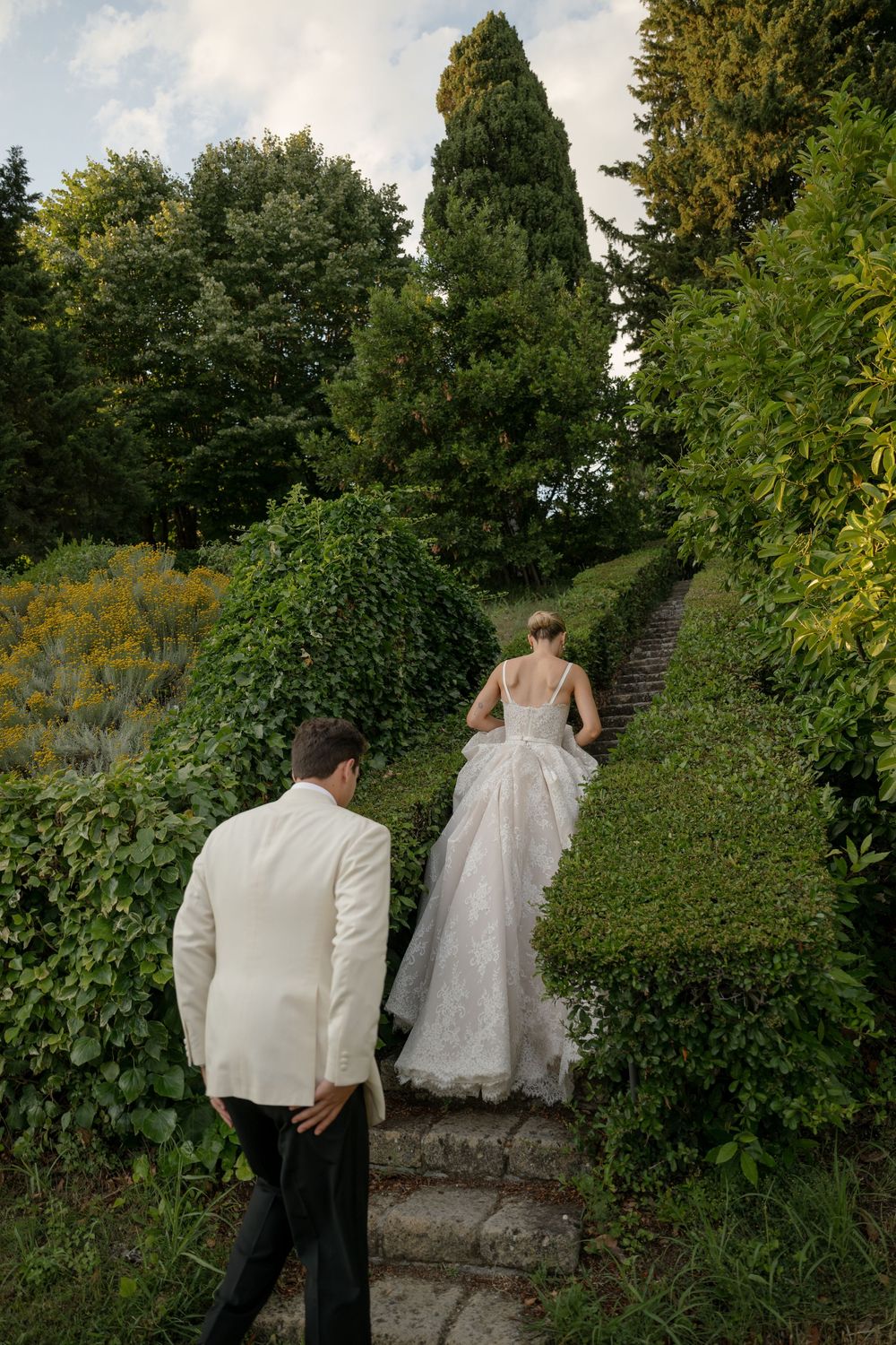 Bride and groom walking up stone garden steps surrounded by lush greenery at an elegant outdoor wedding.