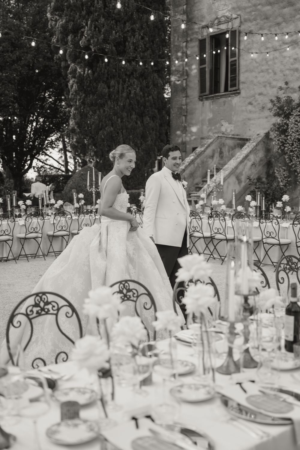 Black and white photo of smiling bride and groom in white tuxedo at elegantly set outdoor reception tables.