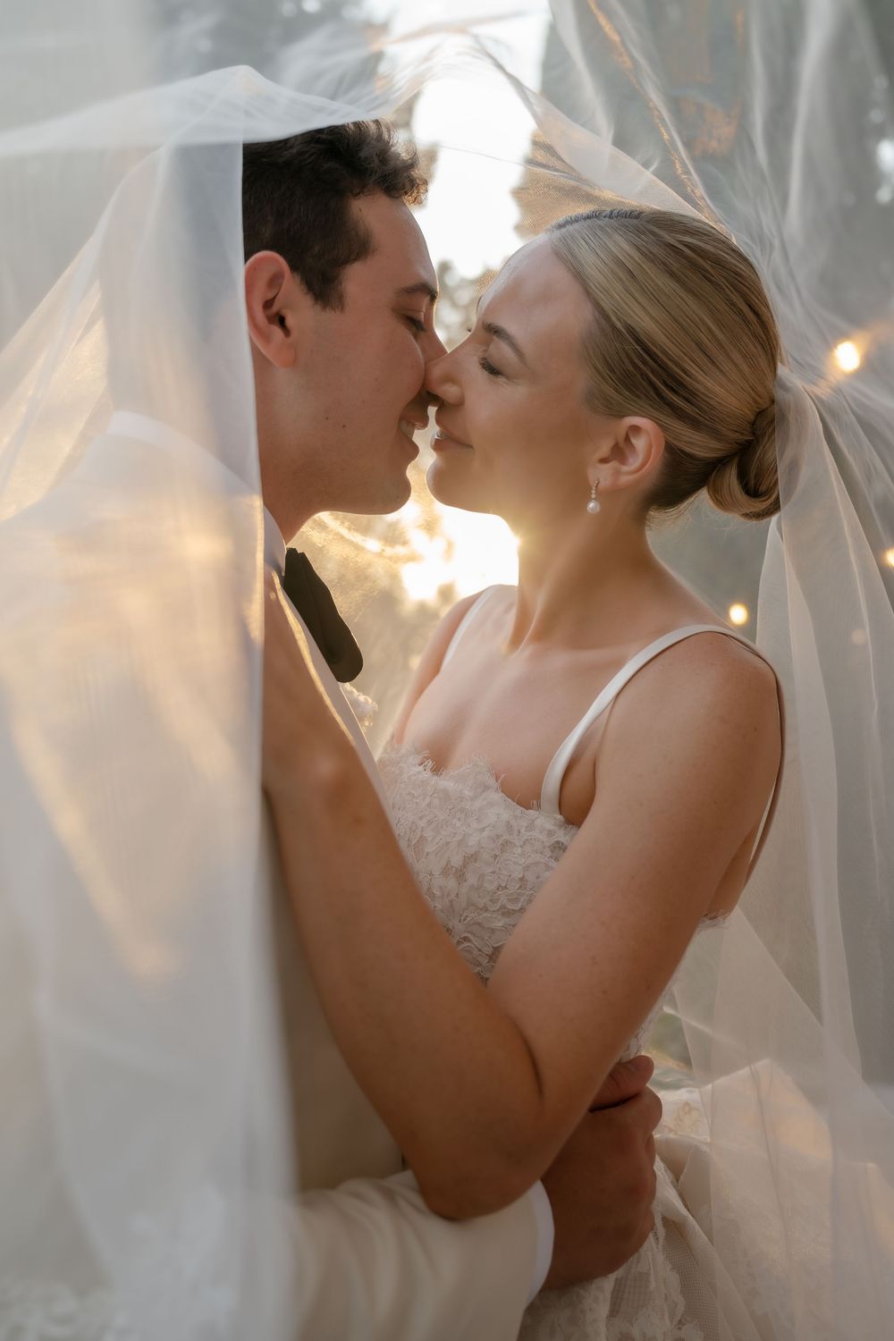 Bride and groom share a romantic kiss under a sheer wedding veil with soft golden backlight.