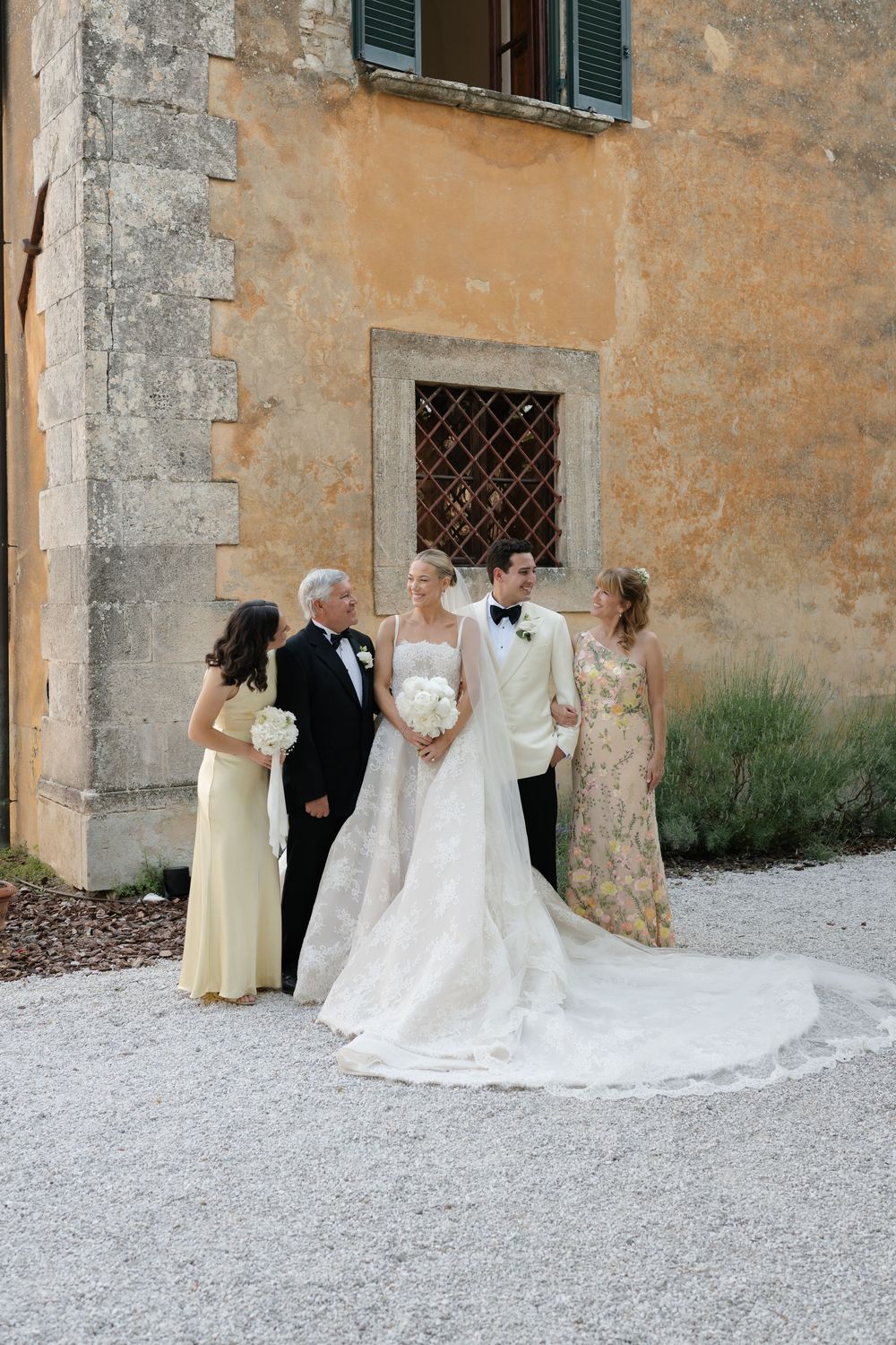 Bride in elegant white gown with long train poses with groom and family outside a rustic Italian villa.
