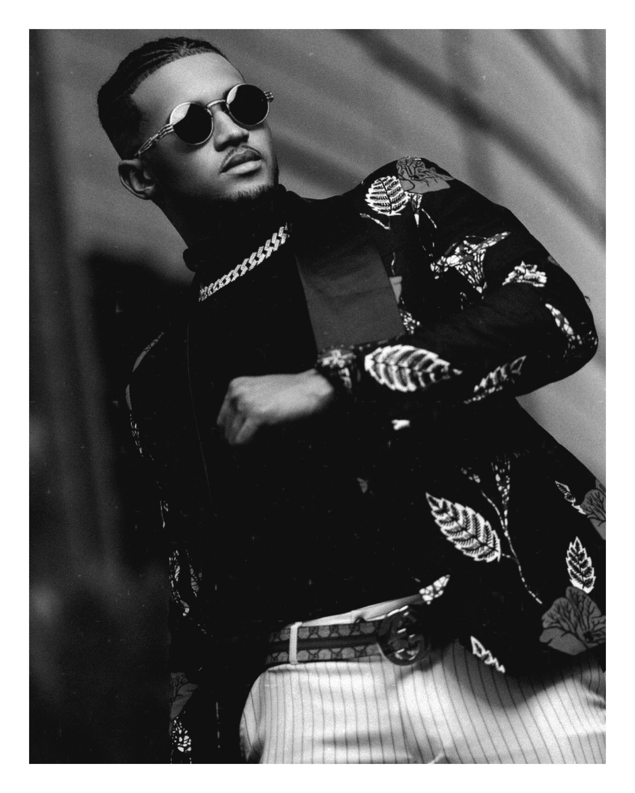 Stylish man in floral blazer and round sunglasses poses confidently in black and white fashion photo.