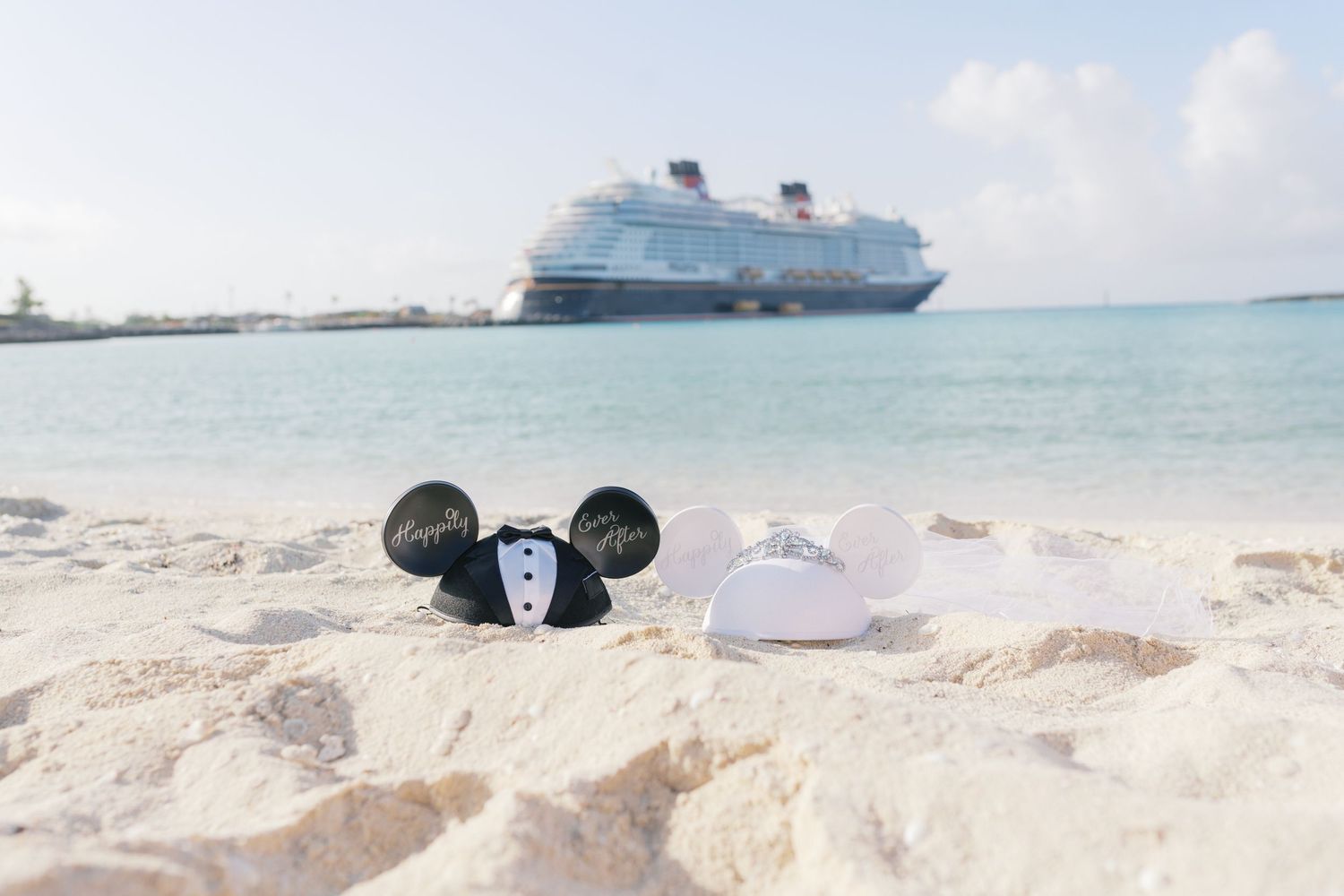 Mickey and Minnie Mouse wedding ear hats in sand with Disney cruise ship in background on tropical beach.