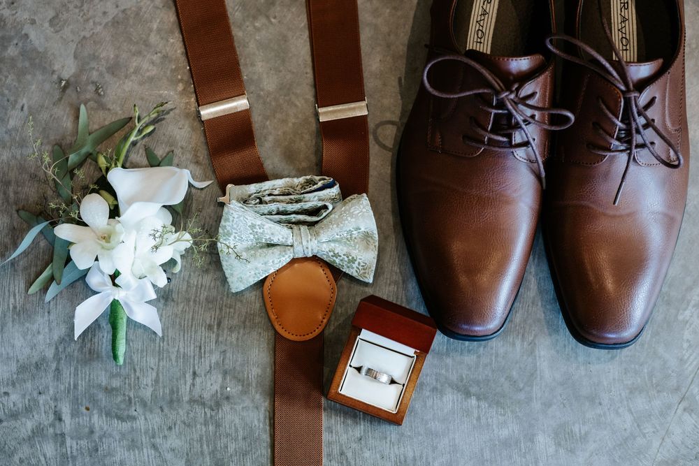 Groom's wedding accessories flat lay with brown leather shoes, suspenders, bow tie, boutonniere, and ring box.