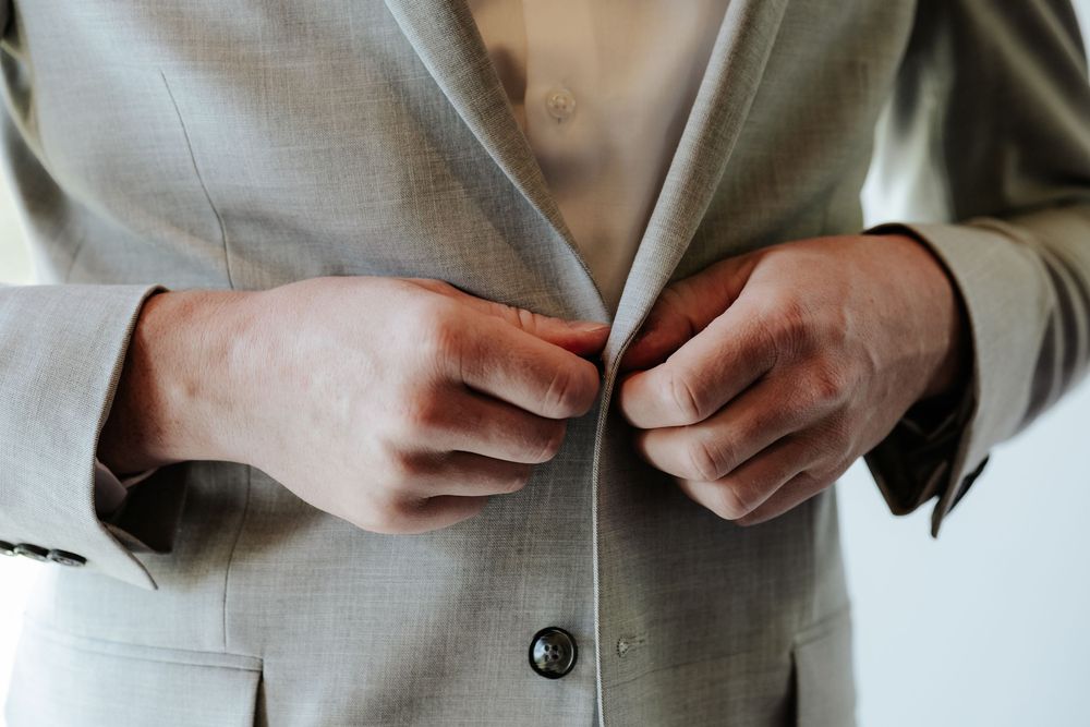 Man buttoning a light gray suit jacket, close-up view of hands fastening buttons on formal menswear.