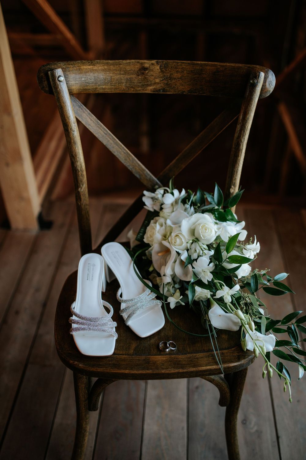 White bridal heels and wedding rings beside a white floral bouquet on a rustic wooden chair.