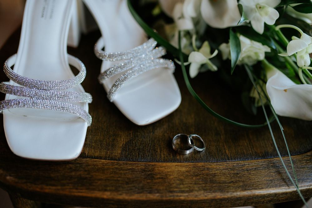 White bridal heels with crystal straps beside wedding rings and white floral bouquet on wooden table.