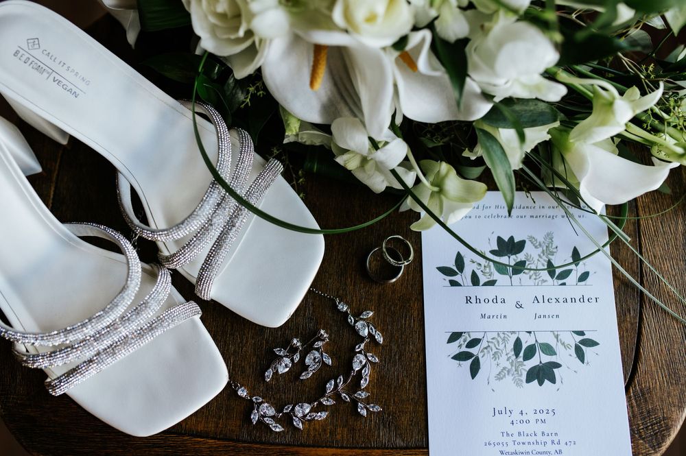 White bridal heels, wedding rings, floral invitation, and white flower bouquet arranged on a wooden surface.