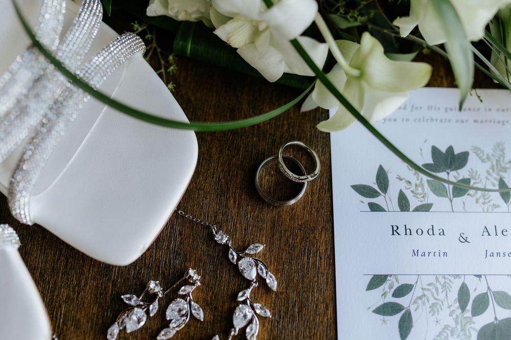 Wedding flat lay with rings, diamond earrings, invitation for Rhoda and Alex, white florals on wood.