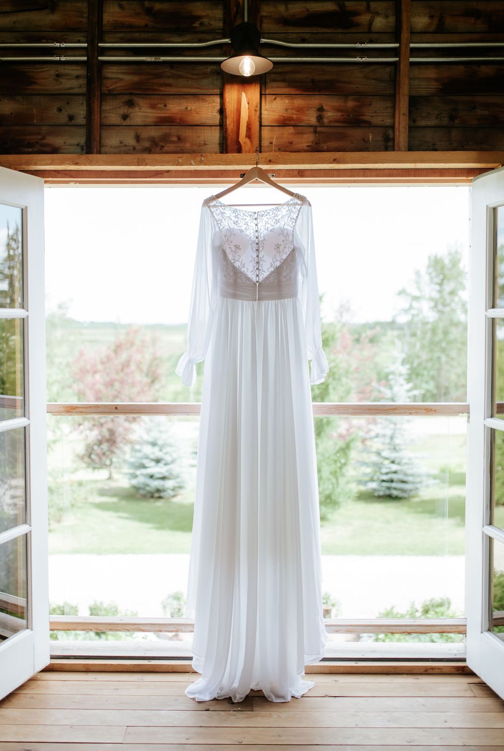 White long-sleeve wedding dress hanging on a wooden hanger in front of open French doors with a garden view.