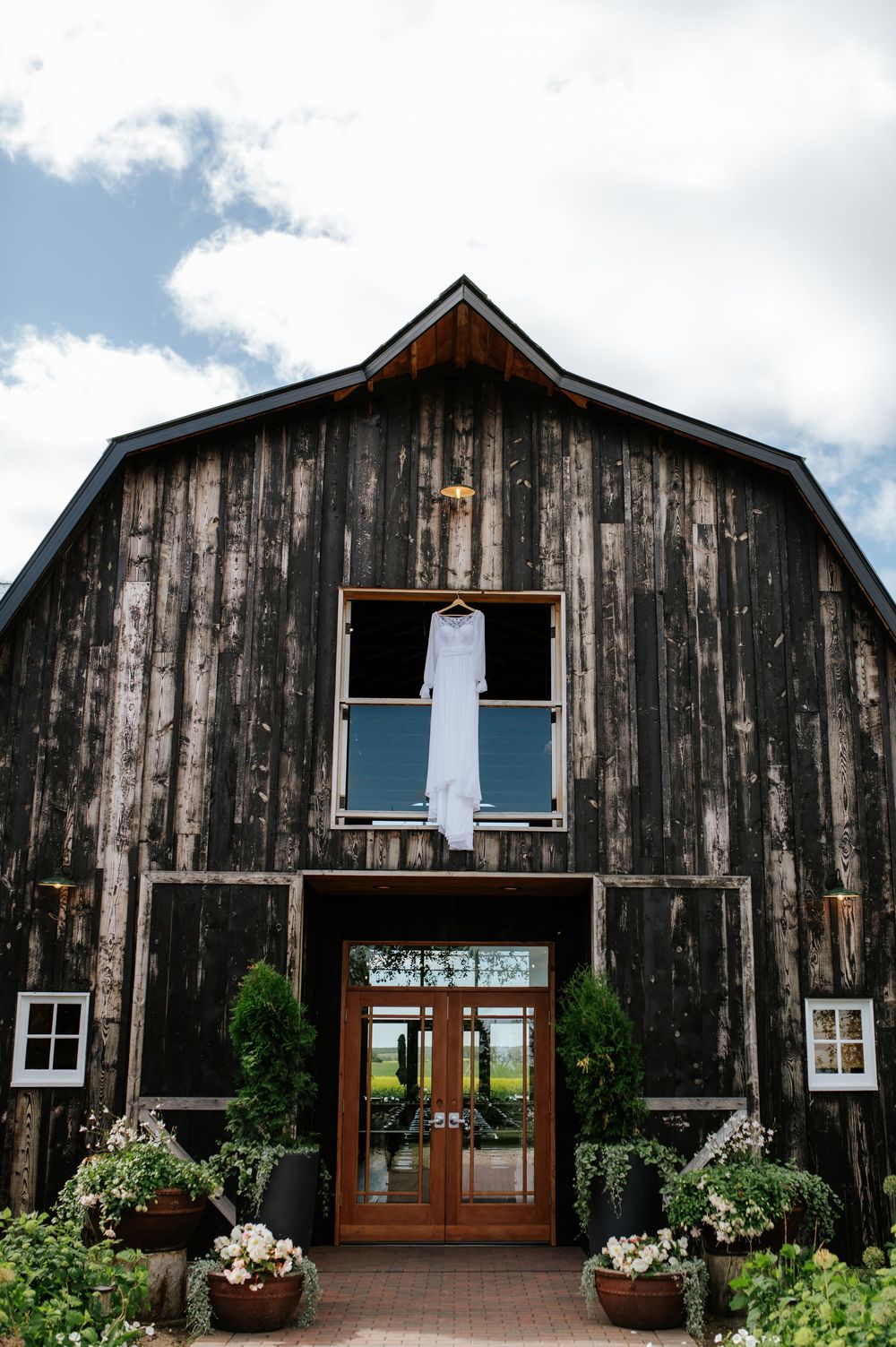 Rustic dark wood barn wedding venue with white gown hanging above entrance, flanked by lush greenery and flower pots.