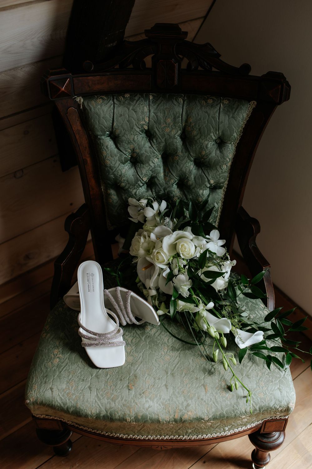 White bridal heels and floral bouquet with roses and greenery resting on a vintage green tufted chair.