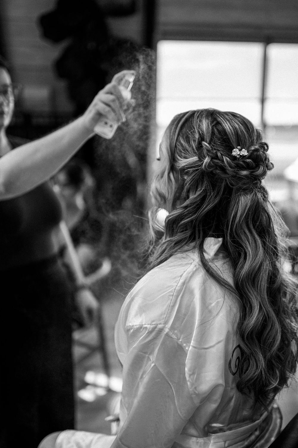 Hairstylist spraying a bride's elegant half-up curled wedding hairstyle with decorative pin.