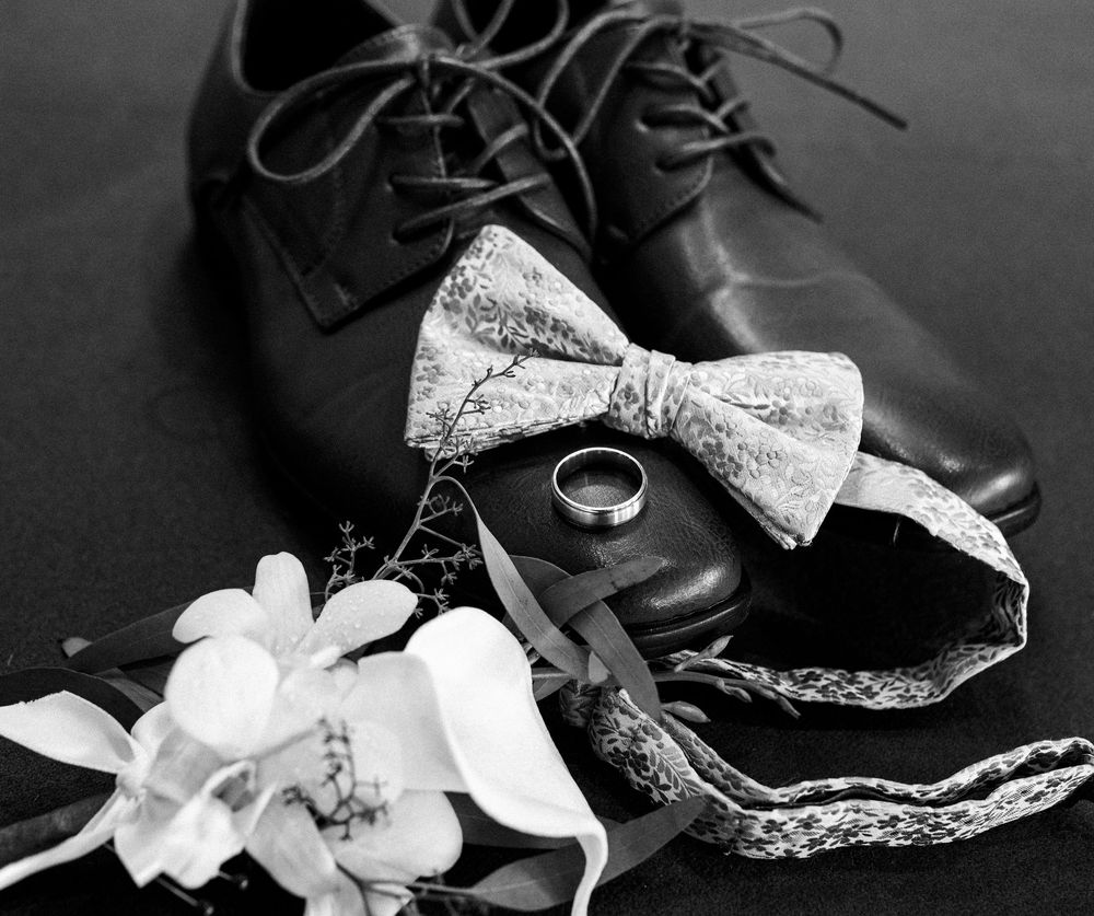 Black and white wedding flat lay with dress shoes, lace bow tie, gold ring, and floral boutonniere on dark background.