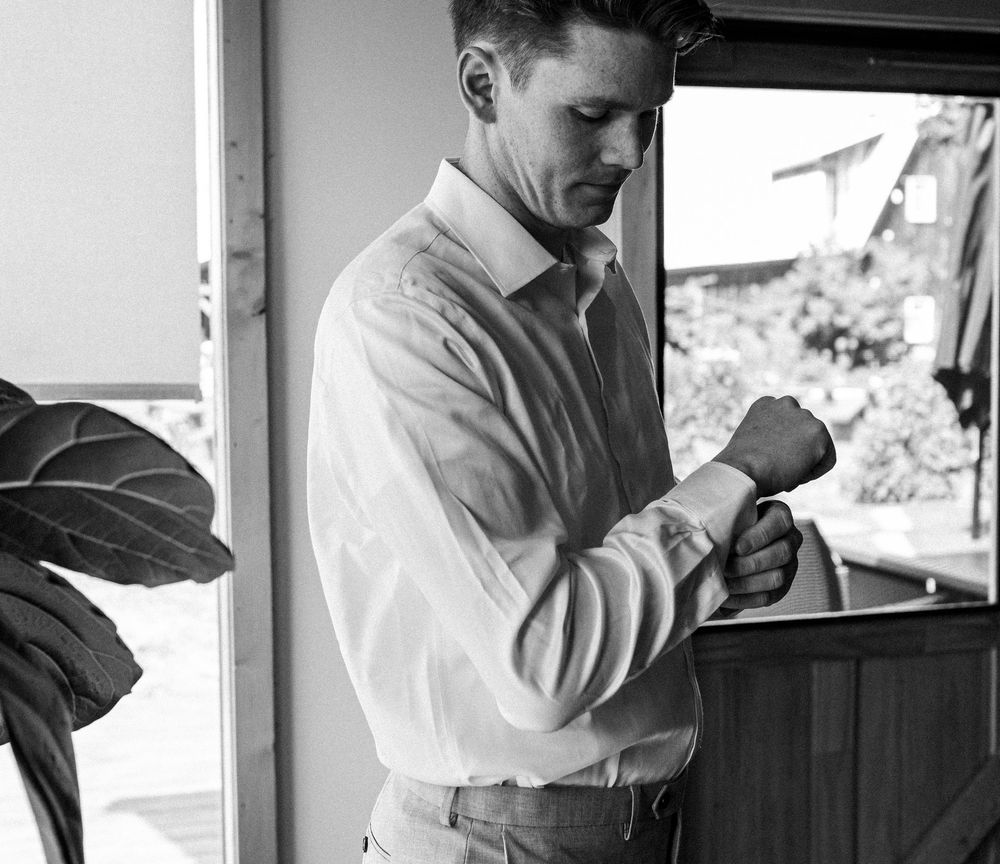 Black and white photo of a man in a white dress shirt adjusting his cufflinks indoors.