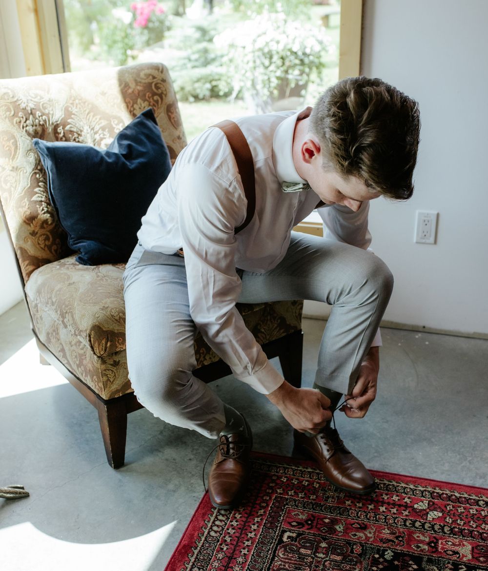A well-dressed groom in white shirt and suspenders ties his brown leather dress shoes while seated in a floral armchair.