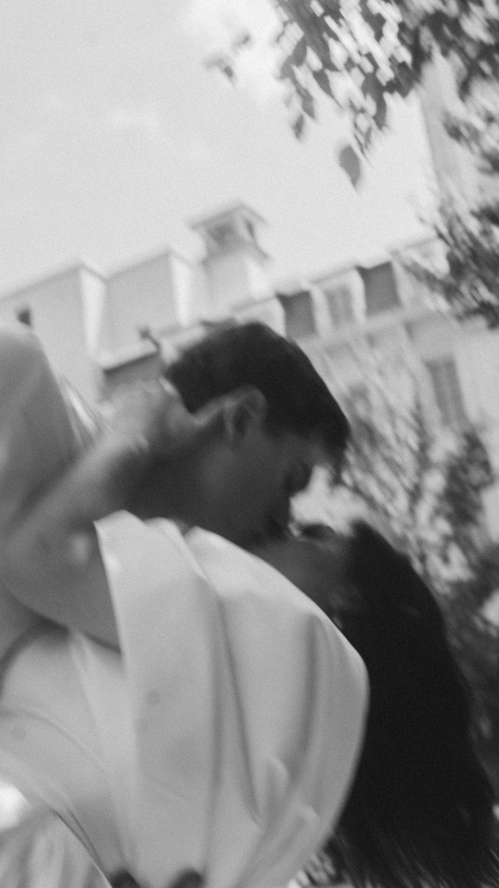Blurry black and white photo of a couple sharing a romantic kiss outdoors near trees and a building.