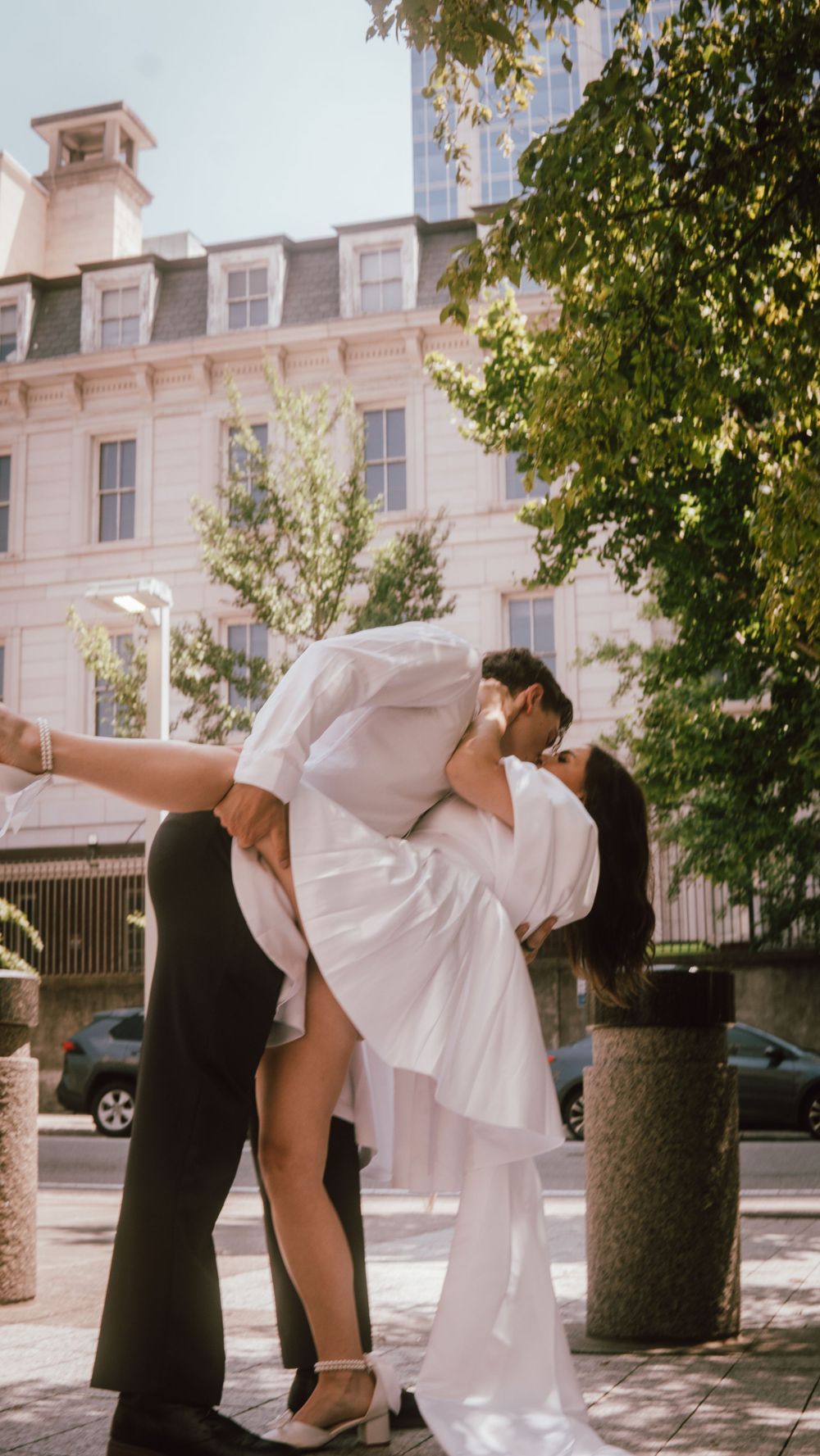 Couple sharing a romantic dip kiss outdoors in wedding attire near a classic building with lush green trees.