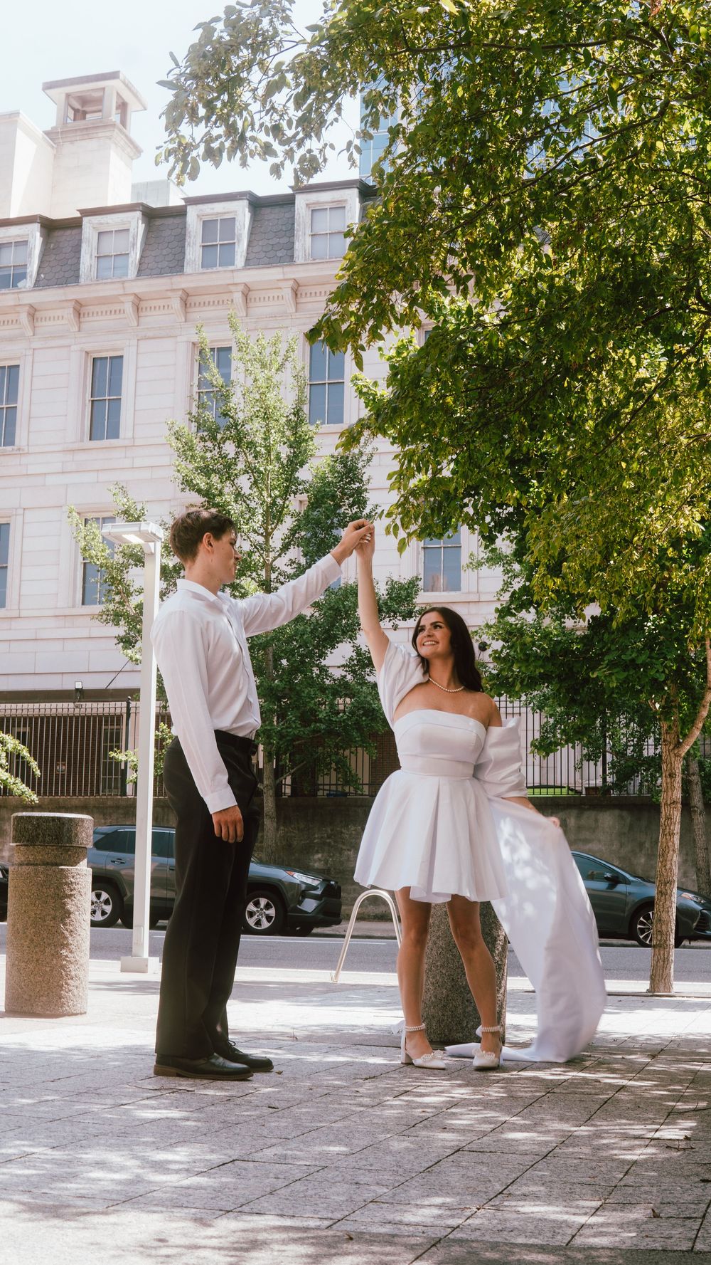 Bride and groom dancing outdoors on a sunny urban street, surrounded by green trees and a classic building.